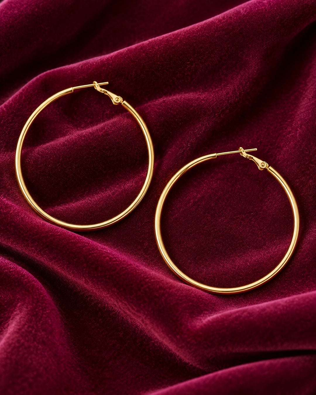 Elli Jewellery large smooth 18ct gold plated hoop earrings resting on a luxurious deep red velvet fabric background.