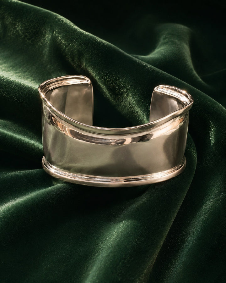 Elli Jewellery rhodium plated minimalist curved bangle bracelet resting on a luxurious dark green velvet fabric.