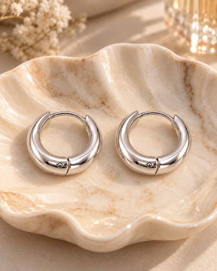 Elli Jewellery rhodium plated minimalist huggie hoop earrings with a polished silver finish resting on a shell dish.