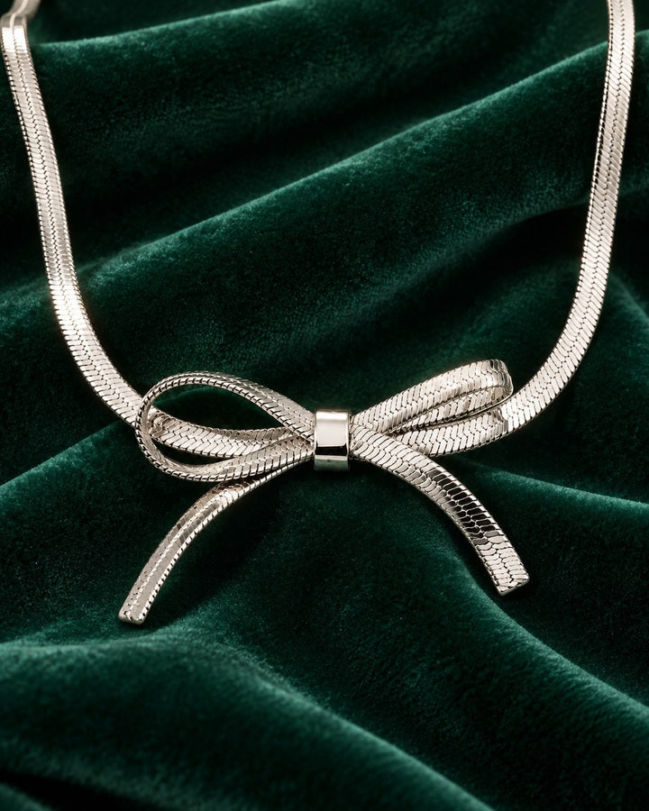 Elli Jewellery rhodium plated snake chain necklace featuring a sculptural bow knot design on dark green velvet.