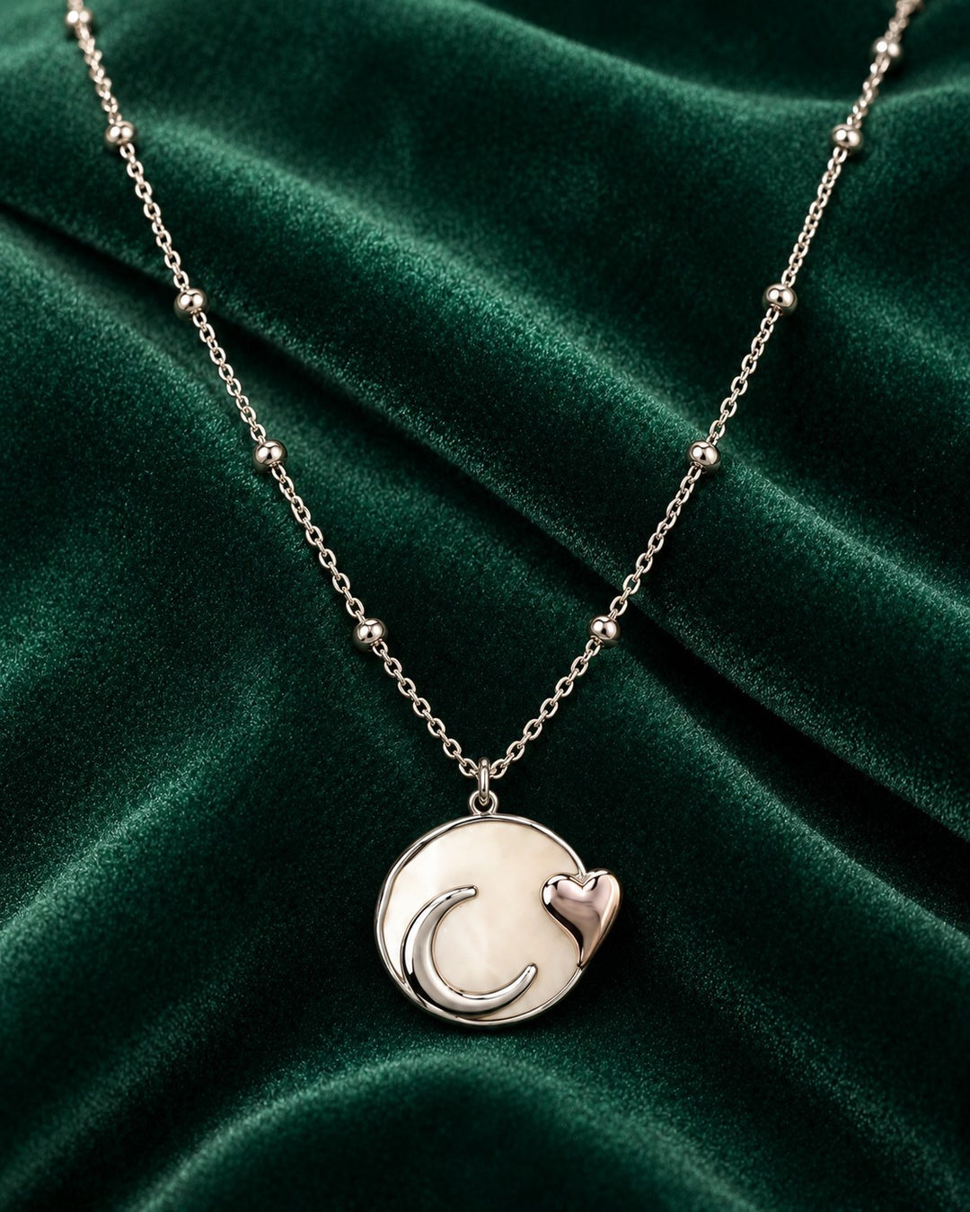Elli Jewellery rhodium plated necklace featuring a moon and heart charm on a beaded chain against green velvet.