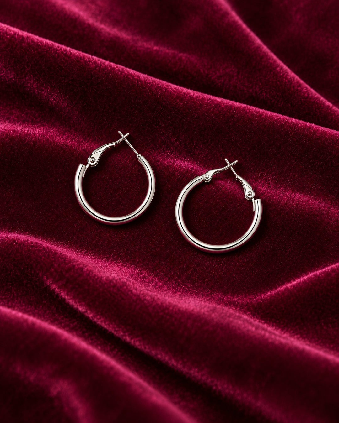 Elli Jewellery rhodium plated hoop earrings with a polished silver finish resting on a deep red velvet fabric.