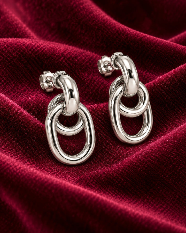 Elli Jewellery rhodium plated drop earrings featuring interlocking oval links on a deep red velvet background.