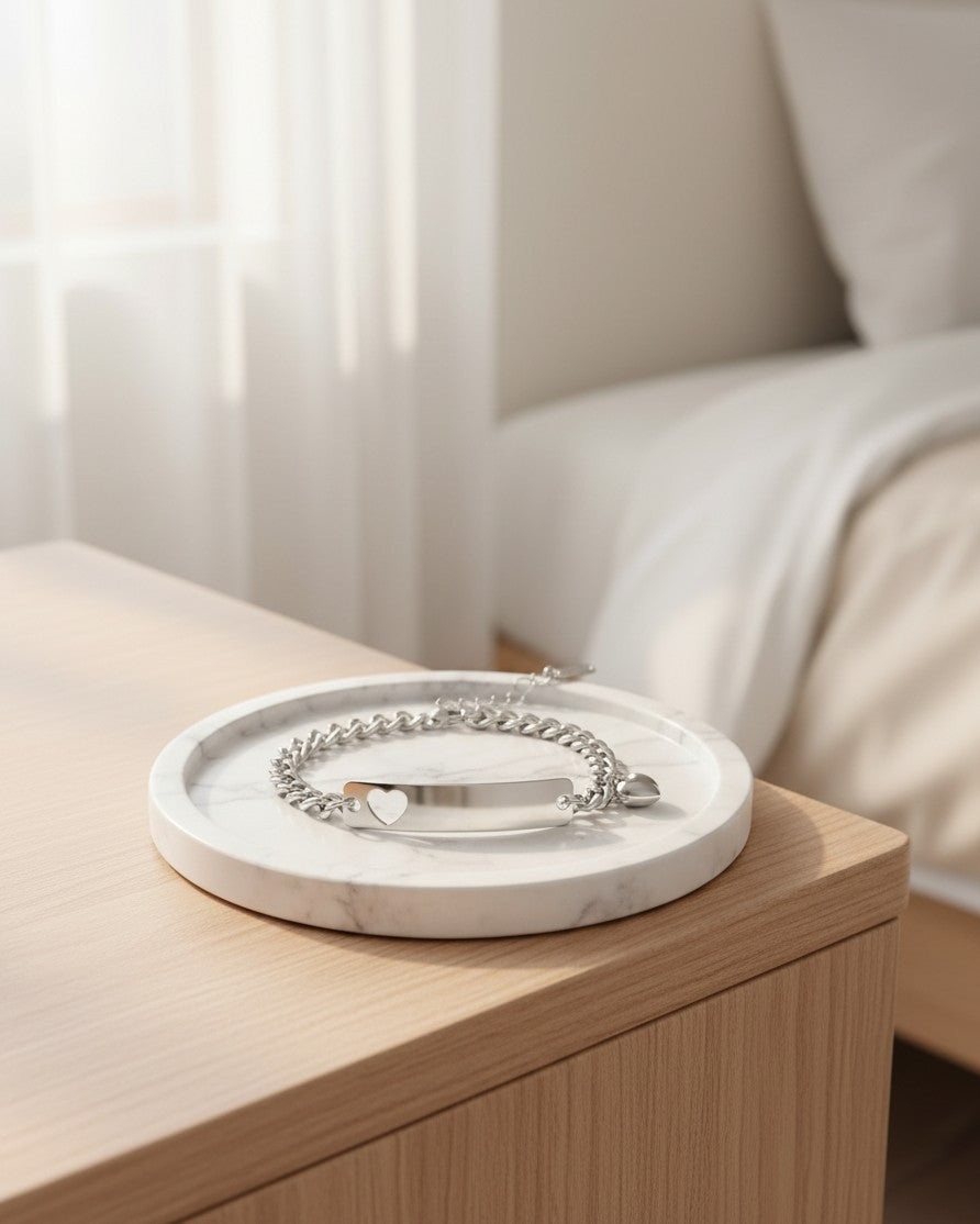 Elli Jewellery silver plated heart ID tag charm bracelet resting on a white marble tray on a wooden bedside table.