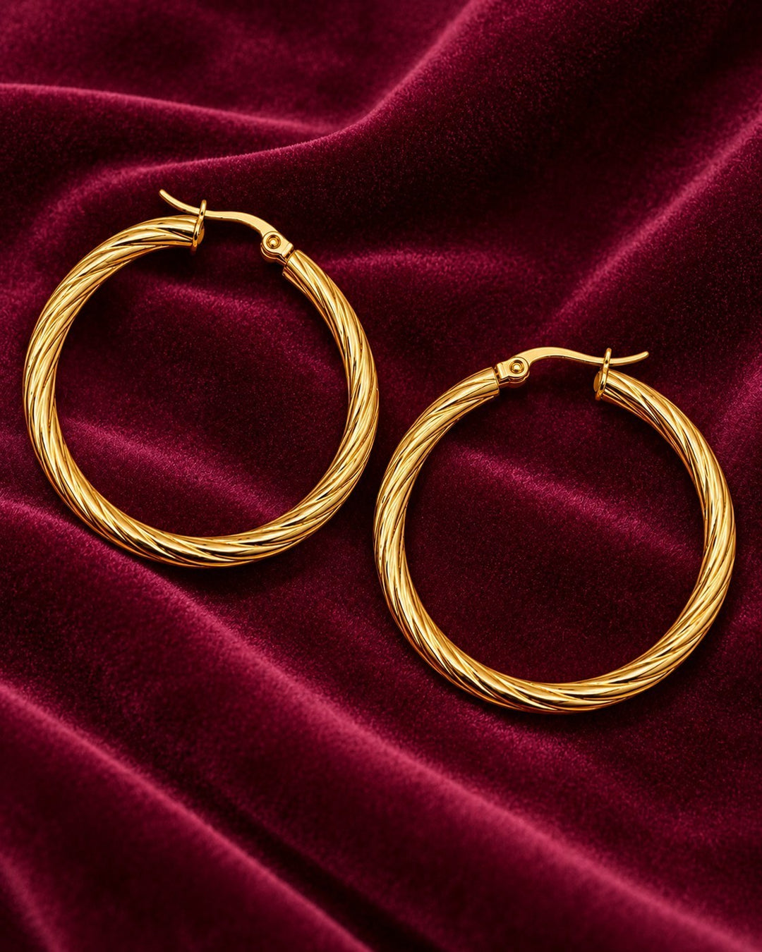 Elli Jewellery 41mm gold-plated twisted hoop earrings resting on a luxurious deep red velvet fabric background.