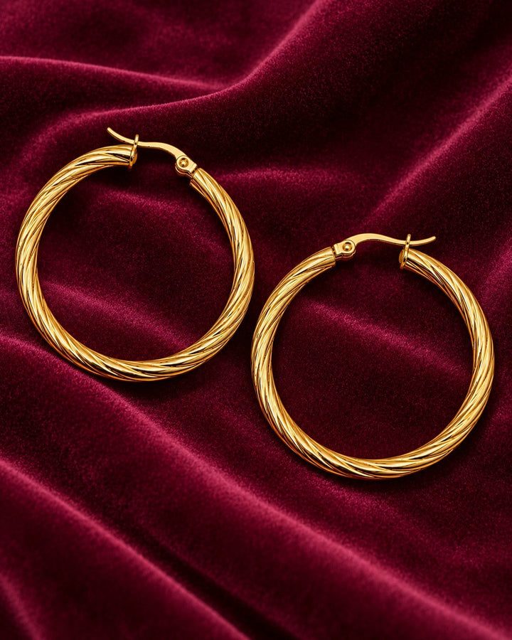 Elli Jewellery 41mm gold-plated twisted hoop earrings resting on a luxurious deep red velvet fabric background.