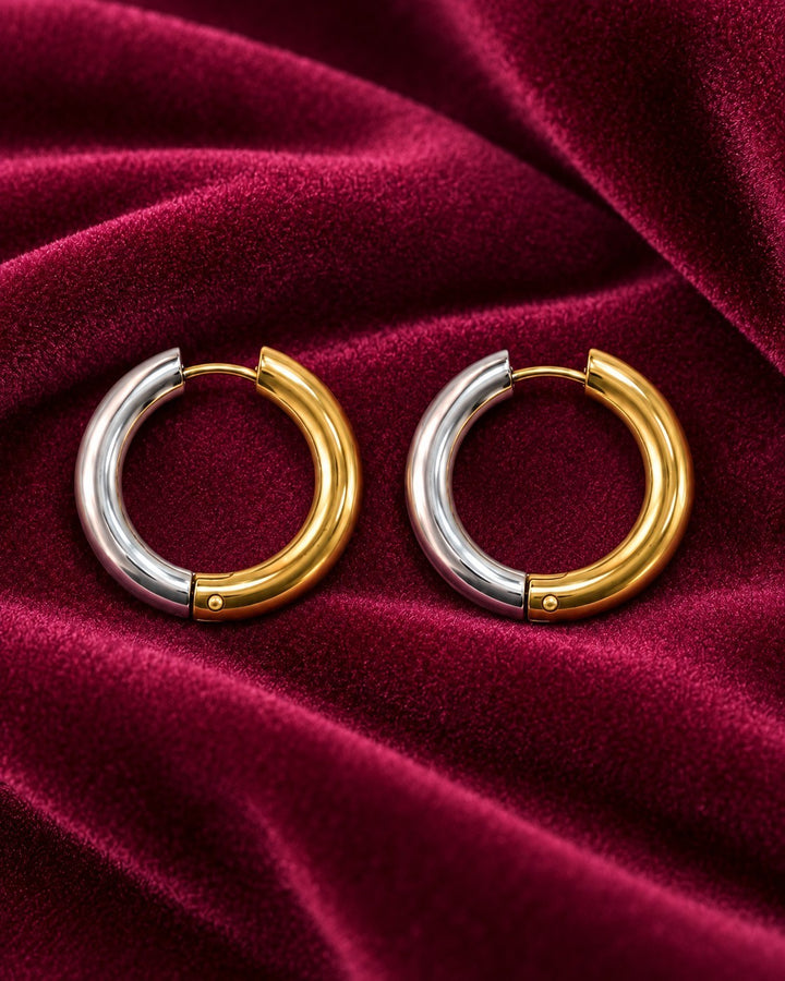 Elli Jewellery two-tone gold and silver 20mm chunky huggie hoop earrings resting on a deep red velvet fabric.