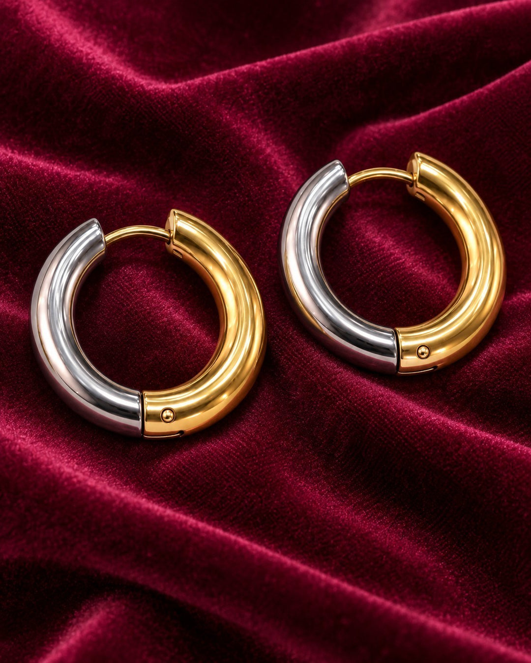 Elli Jewellery two-tone 22mm huggie hoop earrings in gold and silver plating on a deep red velvet background.