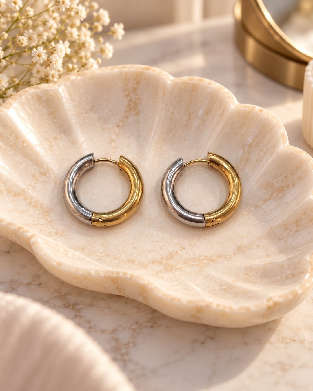 Elli Jewellery two-tone gold and silver 22mm chunky huggie hoop earrings resting in a decorative shell dish.