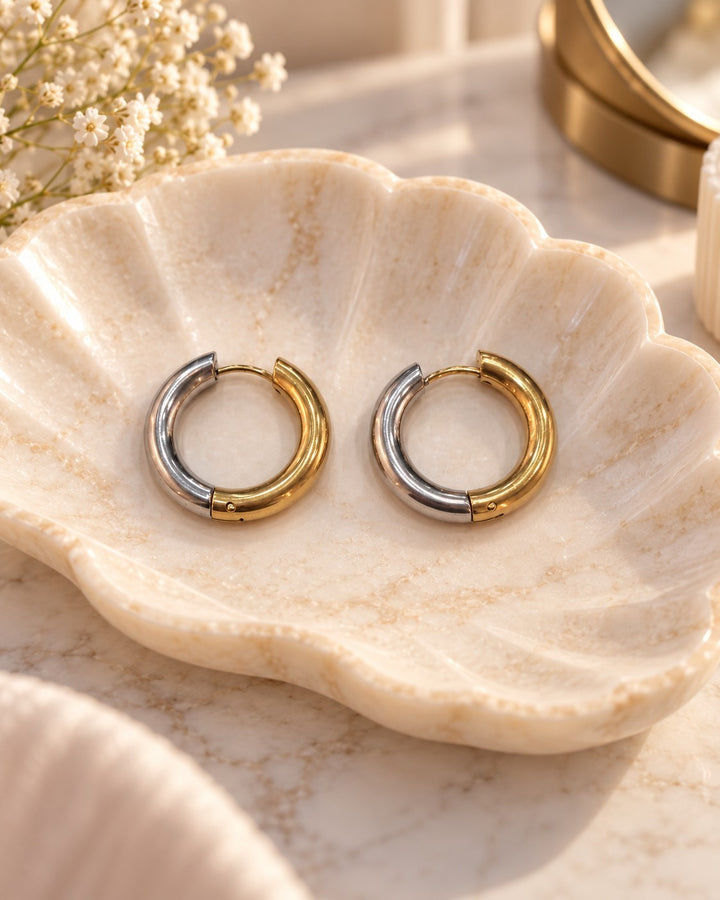 Elli Jewellery two-tone gold and silver 22mm chunky huggie hoop earrings resting in a decorative shell dish.
